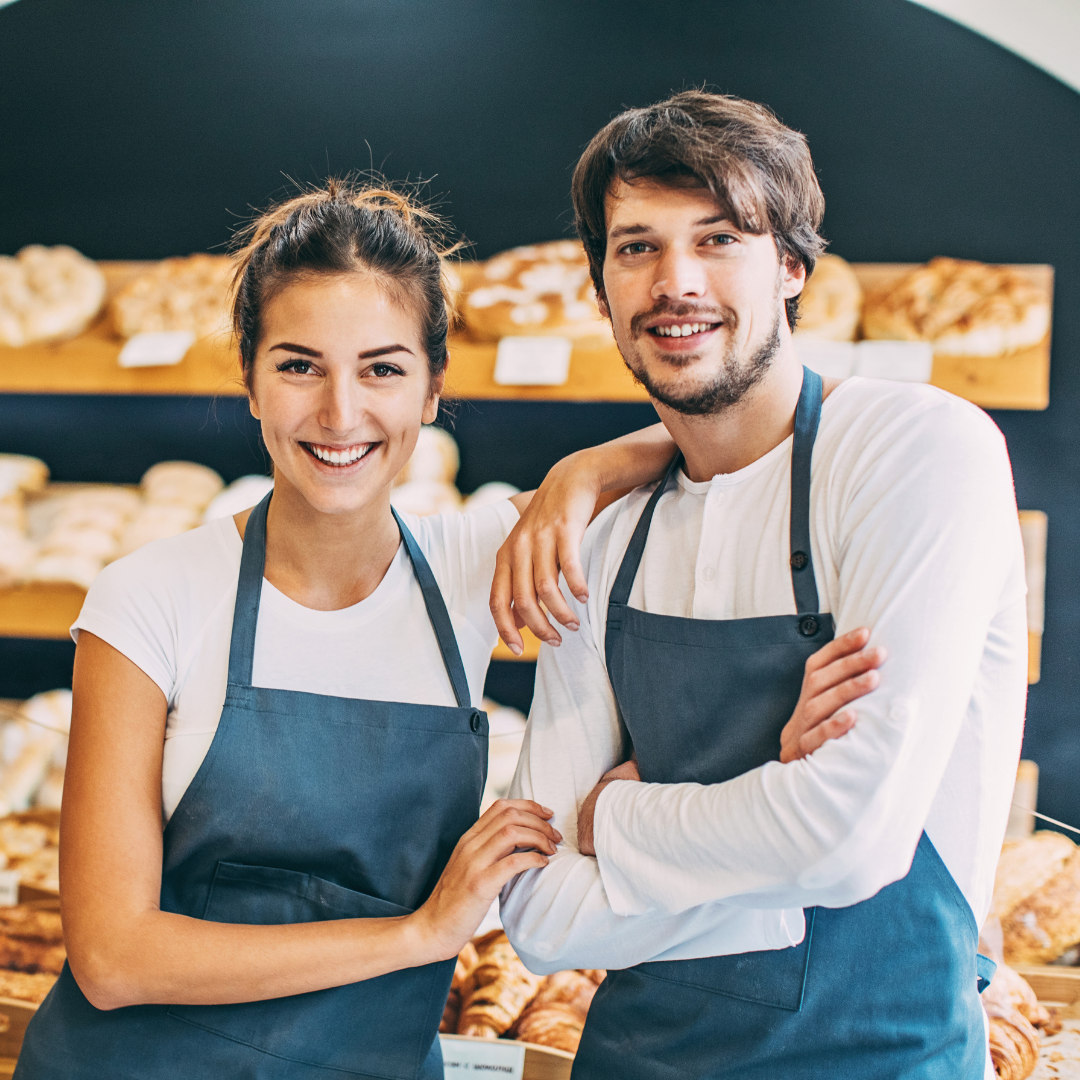 Two bakery staff in blue aprons smile at the camera with bread on shelves behind them.