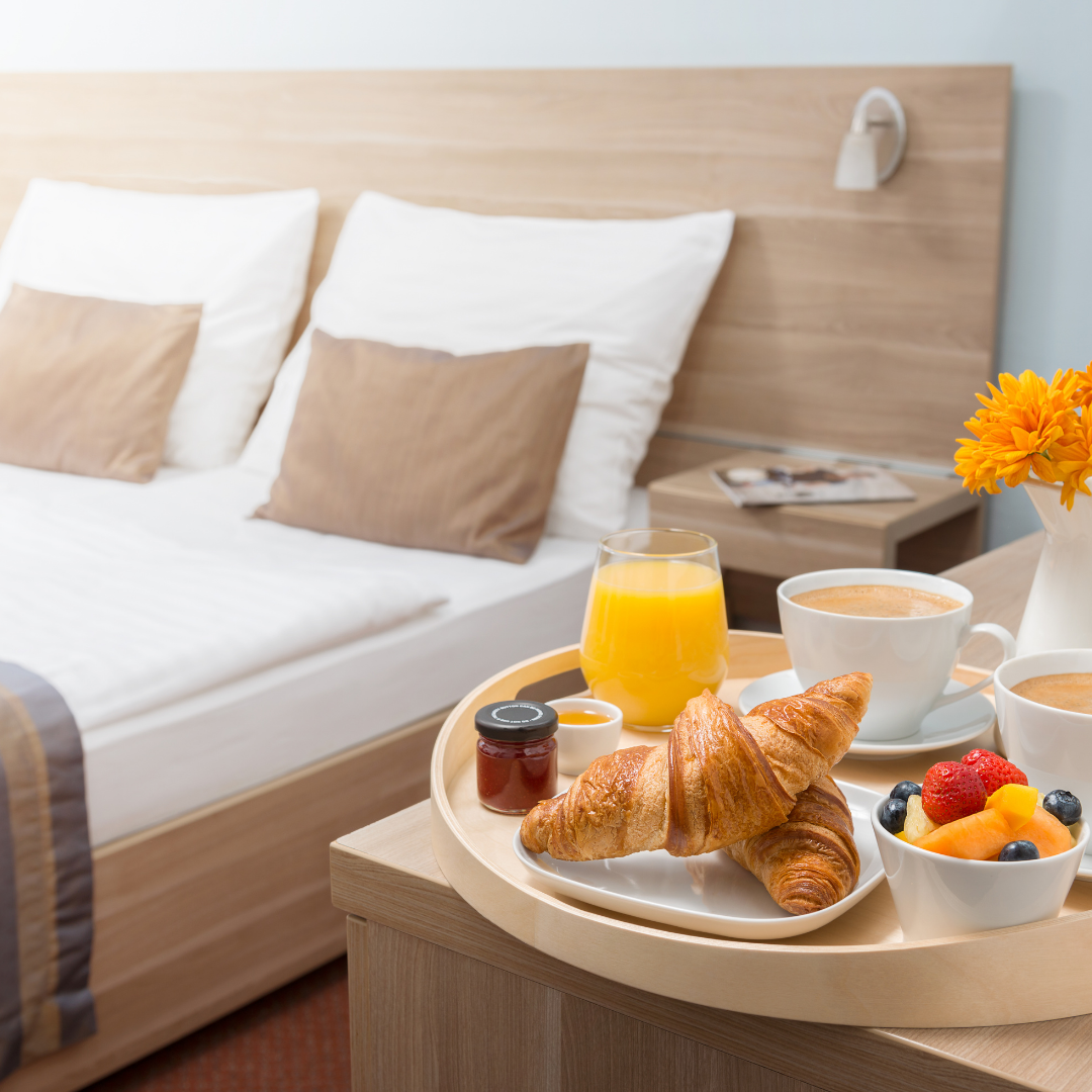Hotel room breakfast tray on a round table beside the bed, with croissants, jam, fruit, orange juice and coffee.