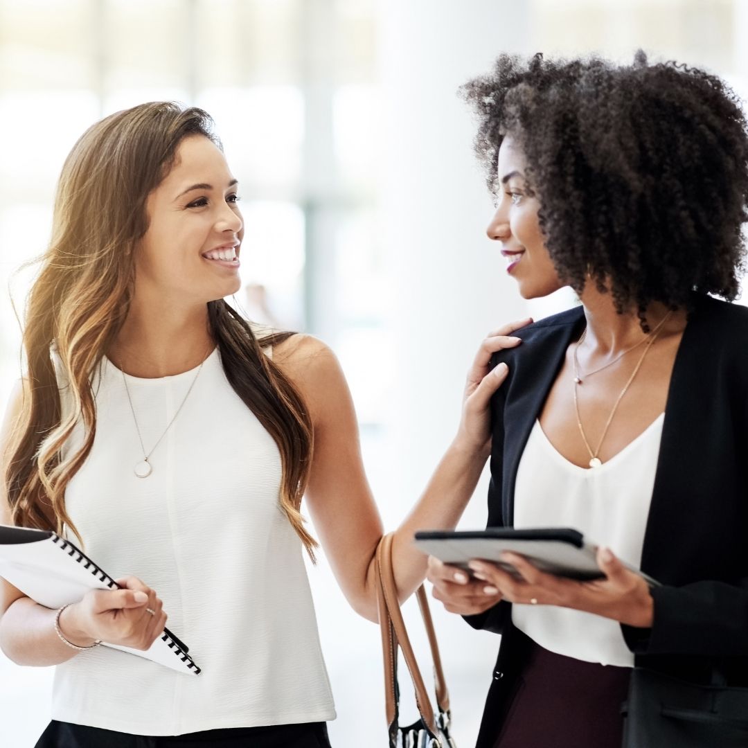 Two professional women walk and chat in a bright office, one holding a notebook and the other a tablet.