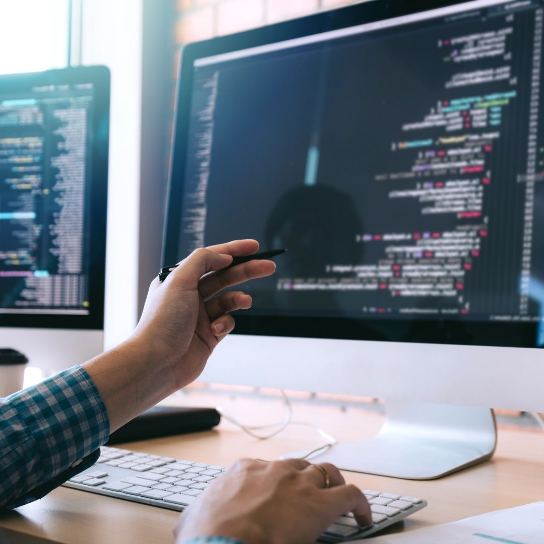 Person coding at a dual-monitor setup, holding a pen while typing on a keyboard.